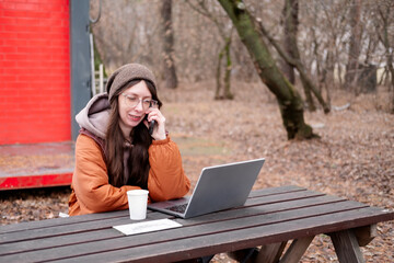A young female freelancer in a brown jacket and hat works remotely at a table in an autumn park, making a business call. Conceptual concepts of freelancing, online business
