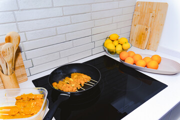 Chicken skewers with marinade cooking on a modern induction hob in a bright kitchen, surrounded by fresh lemons and oranges
