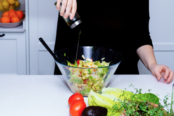 Person preparing healthy green salad, pouring olive oil dressing over lettuce, avocado and tomato in glass bowl on kitchen counter