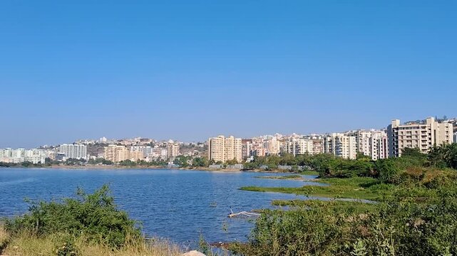 Urban riverside landscape with a calm water body, green vegetation in the foreground, and a modern residential skyline under a clear blue sky, showcasing peaceful city life and natural surroundings.