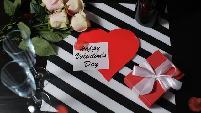 Rose petals fall from above onto a table. The table has a heart-shaped card that says Happy Valentine's Day, a gift box, flowers, and champagne glasses. This setup is perfect for a celebration