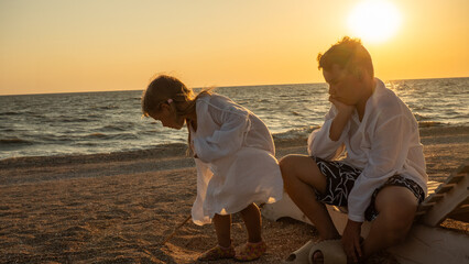 Two children, a boy and a girl, sitting and playing on a sandy beach during a beautiful sunset, sharing a quiet, pensive moment by the ocean as the sun dips below the horizon.