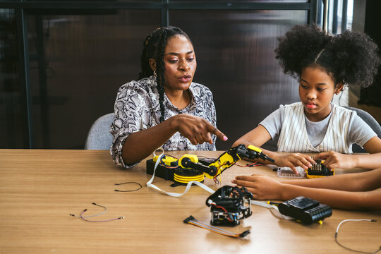 An African teacher and young woman student mentor a curious girl as she assembles a small robot, promoting hands-on STEM learning, teamwork, and coding skills in an inclusive, tech-friendly classroom. - Powered by Adobe