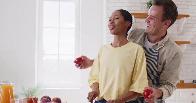 Happy couple cooking romantic breakfast with love and jokes in home kitchen together. Happy young people smile while man in apron making tomato eyes, woman cutting healthy vegetables with knife