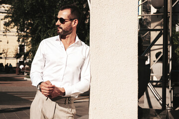 Fototapeta premium Portrait of handsome confident stylish hipster lambersexual model.Modern man dressed in white shirt and trousers. Fashion male posing in the street background in Europe city at sunset. In sunglasses