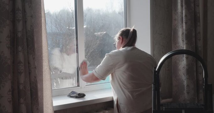 A diligent woman washes dirty windows with a rag and a basin of water during a routine spring cleaning, letting bright sunlight into her cozy home. A woman washes windows in a house.