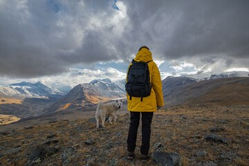 A woman in a yellow jacket and black backpack stands on a mountain road rise, gazing at a valley with snow-dusted peaks and colorful forest. Her large white dog is by her side. © ANDREY