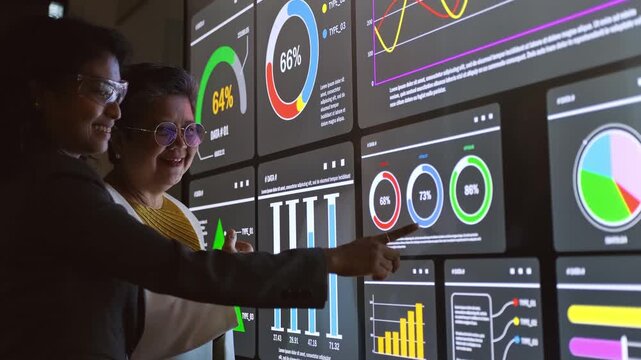 Two professionals analyze data on a large touchscreen, pointing at colorful dashboards and graphs in a dark office, illustrating analytics, teamwork, and decision making.