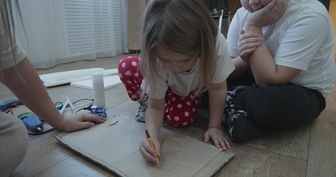 A creative family moment. A little girl, her brother, and her mother are doing crafts together at home, drawing on a piece of cardboard while sitting in the living room. The family is making a craft.