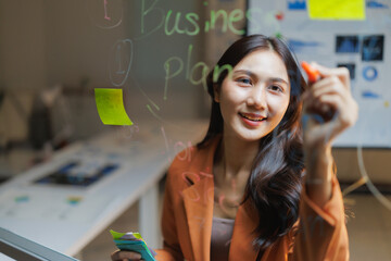 Young Asian businesswoman confidently mapping strategies and ideas on a glass whiteboard in a modern office, smiling while presenting plans for growth, teamwork and innovation