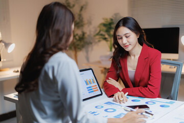 Two businesswomen collaboratively analyzing financial reports and graphs on documents and a digital tablet, working late together to strategize and achieve business goals in an modern office
