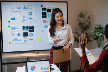 Young Asian businesswoman smiling while presenting financial charts and data on a whiteboard, a colleague raising hand asking a question during a corporate meeting in an office setting