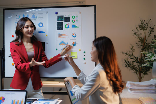 Two businesswomen collaborating and discussing financial data charts on a whiteboard in an office, illustrating teamwork, corporate planning, and successful strategy development - Powered by Adobe