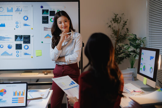 Young Asian business women collaborating on a business project, discussing market research data and financial planning in a modern office environment during an evening meeting - Powered by Adobe
