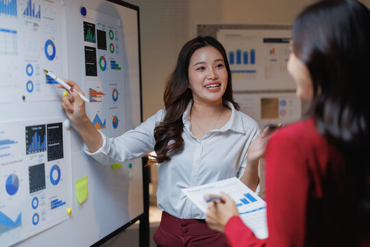Businesswoman explaining marketing strategy to colleague during corporate presentation, pointing to charts and graphs on whiteboard while discussing data, planning and teamwork