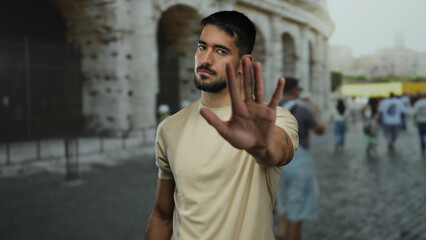 Fototapeta premium Young man standing outside near the roman coliseum in italy with a hand gesture, blending hispanic beauty with historic city charm in a vibrant street atmosphere.