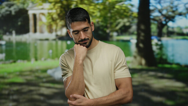 Pensive man outdoors in park setting with thoughtful expression wearing casual beige shirt surrounded by lush trees and peaceful lake under vibrant sunlight