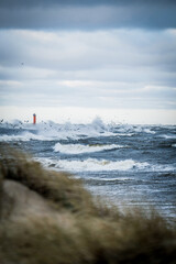 Fototapeta premium Stormy sea with strong waves crashing under a dramatic cloudy sky. A red navigation beacon stands in the distance while seabirds fly above the rough water. Windy coastal landscape