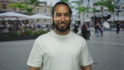 Fototapeta premium Man face winks one eye while smiling and showing beard on a street terrace restaurant patio near outdoor diners in a casual white tshirt; friendly ease.