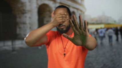 Man with hand over eyes and raised palm to stop, wearing bright orange shirt and necklace at coliseum building tourist area; privacy avoidance distress.
