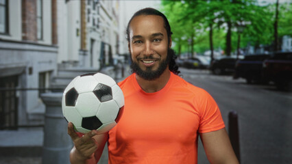 Man in bright orange shirt holds a soccer ball with his fingertips while smiling and balancing on a city street with parked cars and trees; confidence skill.