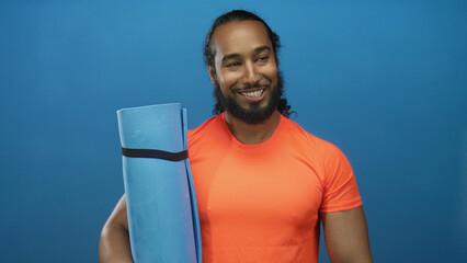Man holding a blue yoga mat, smiling with visible arm and wearing orange shirt in blue studio; confidence wellness.