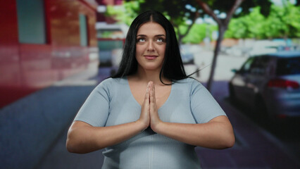 Woman meditating outdoors in a city street poses peacefully wearing casual attire with hands together, embracing tranquility surrounded by urban trees, cars, and buildings.