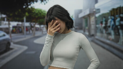 Fototapeta premium Young hispanic woman in cropped top covers eyes with hand against bright sun glare while standing on busy urban street; embarrassment.