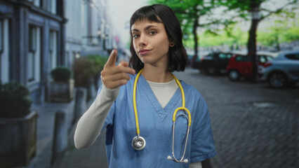 Woman nurse in blue scrubs with yellow stethoscope points finger toward camera on a cobblestone...