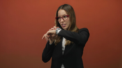 Young woman in black blazer checking smartwatch and pointing to wrist with bare hands in orange studio; time pressure impatience.
