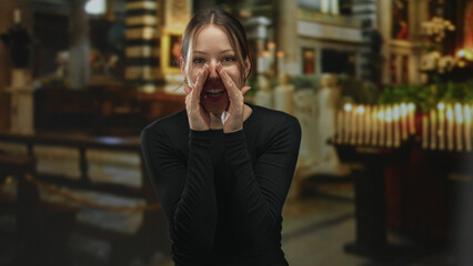 Woman with hands cupped to mouth calling aloud in a catholic church building by lit votive candles and wooden pews, facing forward; joyful praise.