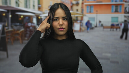 Fototapeta premium Woman in black shirt pointing and thinking on an outdoor street with blurred background of colorful buildings and people walking.