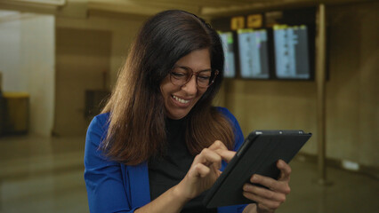 Woman in blue blazer and glasses holds tablet and pumps fist in crowded airport terminal while smiling broadly with excitement  victory. © Krakenimages.com