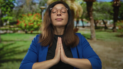 Woman with eyes closed holds hands pressed together in prayer gesture in green park surrounded by...