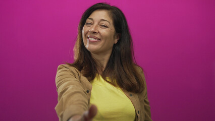 Fototapeta premium Woman extends hand toward camera in pink studio wearing yellow shirt; greeting trust connection welcome.