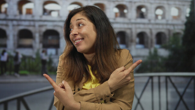Smiling hispanic woman pointing fingers to sides at sunlit ancient roman coliseum building; amusement.