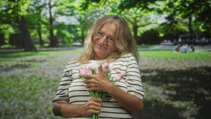 Obraz premium Woman holding pink tulip bouquet close to chest, hands clasping stems against sunlit forest and green grass with distant picnic bench; contentment nostalgia.