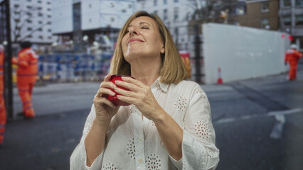 Woman holding red cup near nose while standing on street at construction site wearing white blouse and focusing on drink aroma; serenity.