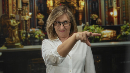 Woman with glasses points finger to eyes while standing before ornate altar in church building;...