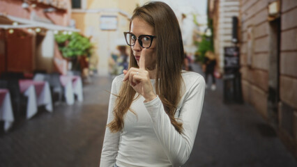 Young hispanic woman wearing glasses points finger at outdoor terrace restaurant on cobblestone street; confidence assertiveness.