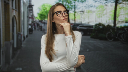 Fototapeta premium Woman in white top wearing glasses with hand on chin and crossed arms on street, looking up and pondering; pensive doubt decision.