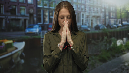 Woman with hands pressed together in prayer, closed eyes, red string bracelet and wristwatch, standing on an amsterdam street by a canal; serenity.