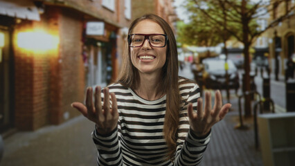 Woman with glasses beckons with both hands on a busy city street, smiling broadly; welcoming playfulness casual.