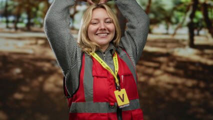 Woman volunteer making heart symbol in park wearing red vest and grey sweater smiling outdoors with sunlight filtering through trees.
