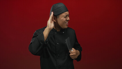 Young hispanic man chef in black uniform holding a cleaver while cupping his ear to listen in a red...