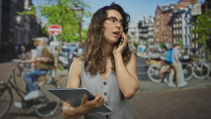 Woman wearing glasses holding clipboard and smartphone to ear on busy city street; multitasking concentration.