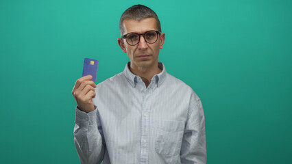 Man with glasses holding credit card against green background looks thoughtful about financial...