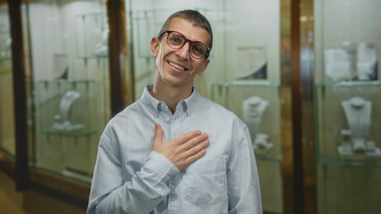Obraz premium Man smiling indoors near jewelry showcase wearing glasses and striped shirt with one hand on chest in a store setting expressing satisfaction and positive emotion.