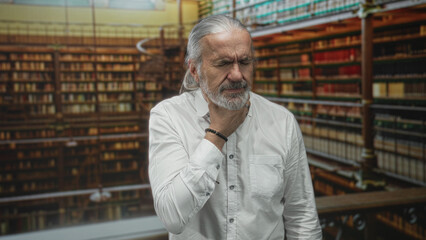 Fototapeta premium Man clutching throat and wincing amid crowded library stacks in an old building, hoary long hair and white shirt visible; choking distress.