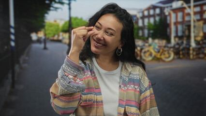 Woman pinching fingers near nose on street while squinting and smiling at camera, wearing patterned jacket; playful teasing.